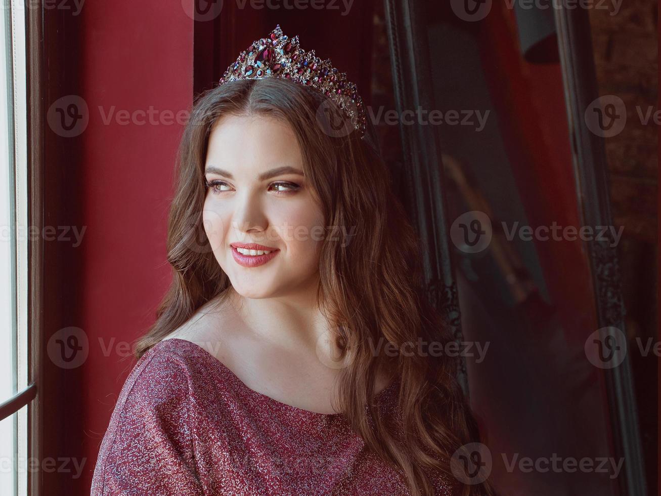 portrait of beautiful young pretty cute woman princess in long purple queen's dress and crown, with long hair and make up indoor in loft interior sitting by the window photo