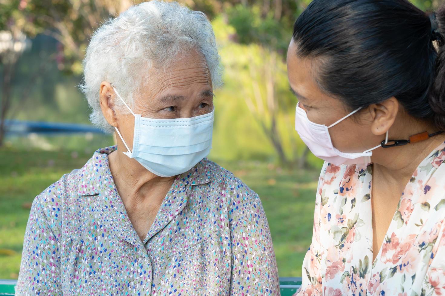 Caregiver help and talk with Asian senior or elderly old lady woman wearing a face mask for protect safety infection Covid-19 Coronavirus in park. photo