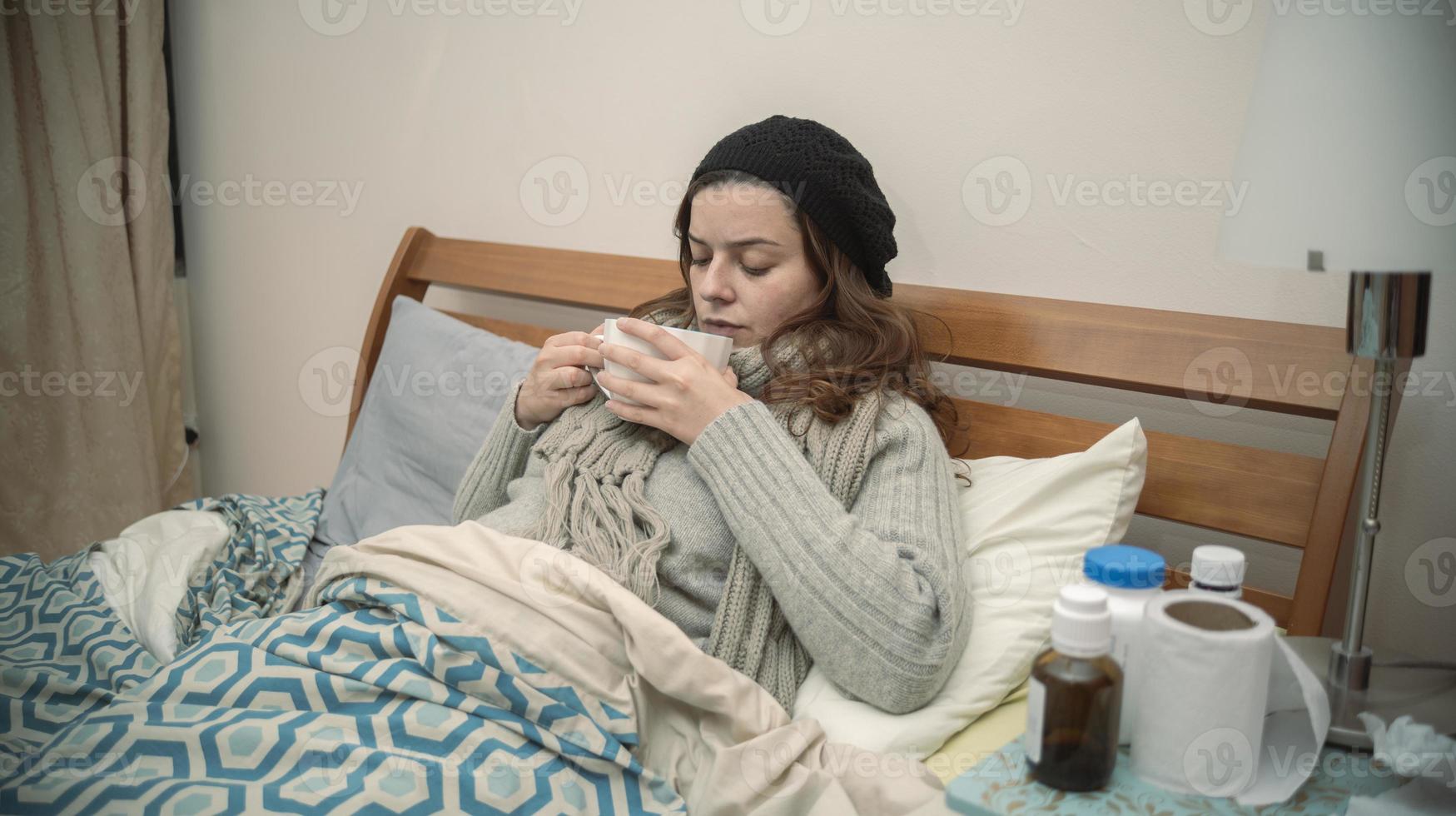 Hispanic woman lying alone in her bed well wrapped up, sick with the flu, drinking a cup of tea photo