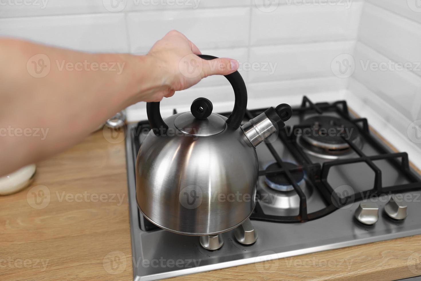 mano de hombre sosteniendo un hervidor metálico en la cocina. La tetera utiliza agua caliente para hervir bebidas como té, café, leche en polvo u otras. foto
