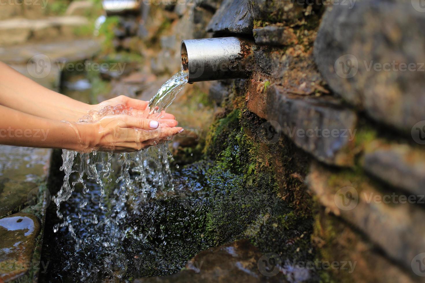 Woman collect pure water in hand palm from the source in the wall, hold and drink it. Female hand scooping spring water from the stone in forest photo