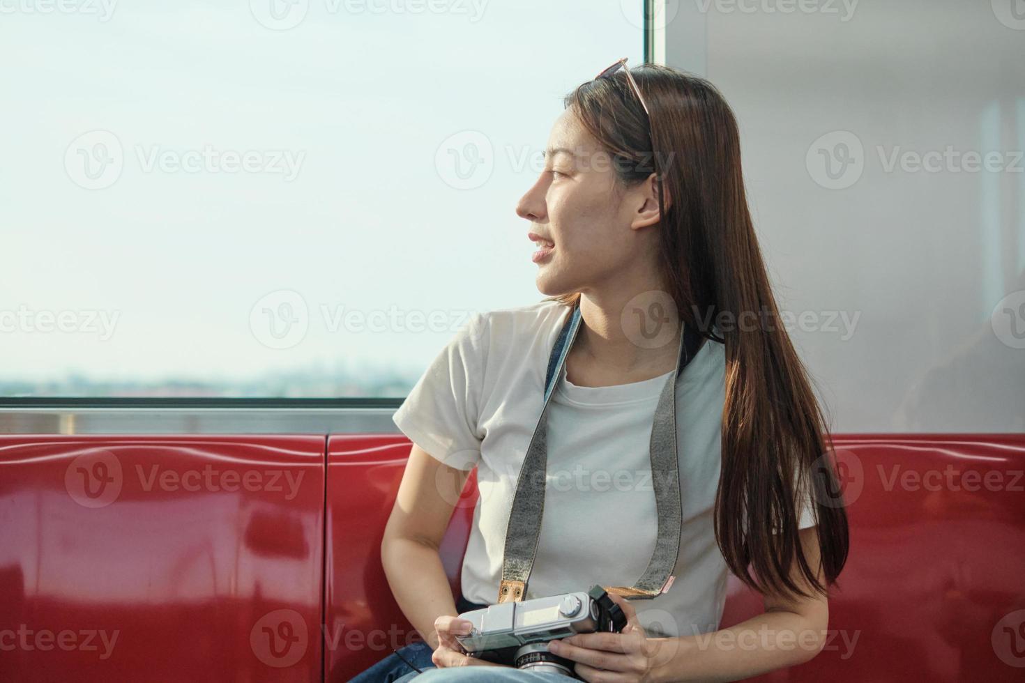 Beautiful Asian female tourist with camera sitting, looking travel location and sightseeing urban window view, public train transport, city lifestyle journey by railway, happy vacation in Thailand. photo