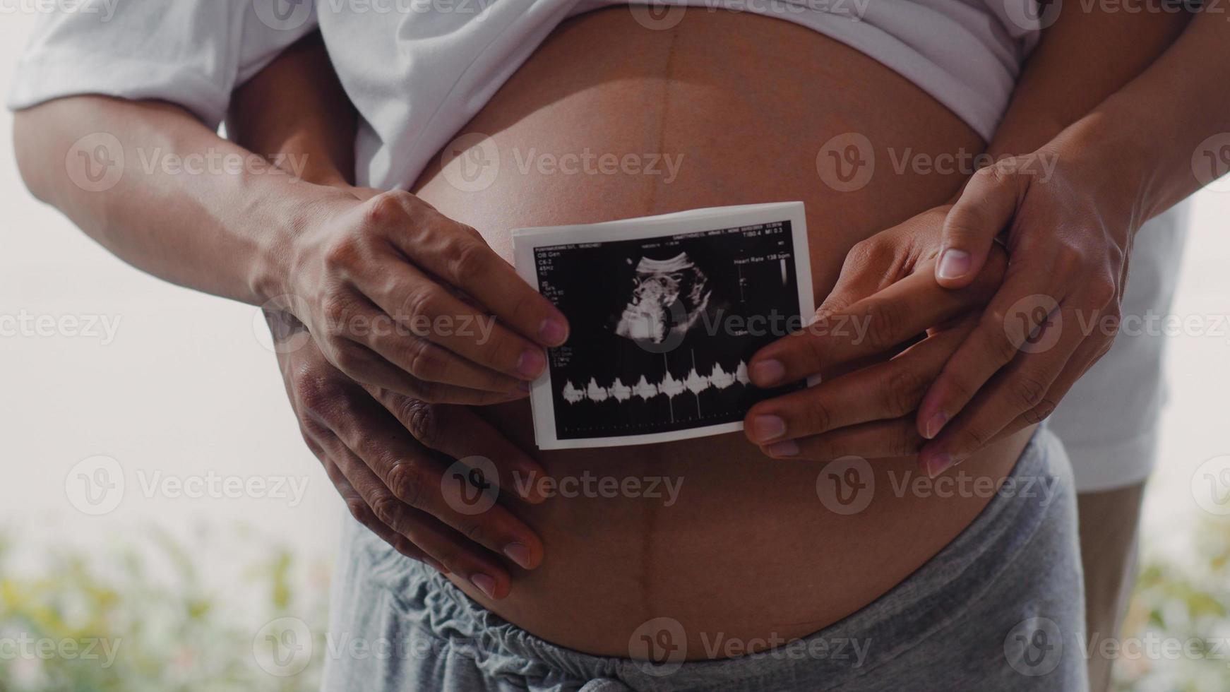 joven pareja asiática embarazada muestra y mira una foto de ultrasonido bebé en el vientre. mamá y papá se sienten felices sonriendo en paz mientras cuidan al niño acostado cerca de la ventana en la sala de estar en el concepto de casa.