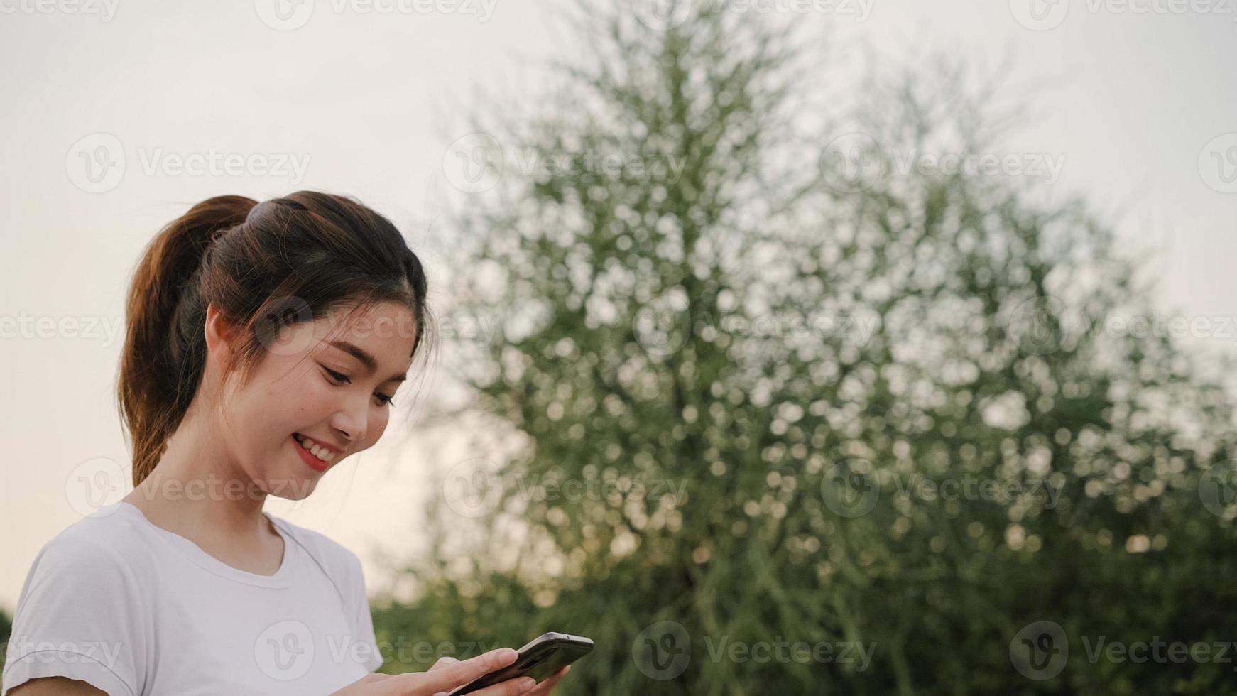 Cheerful Asian tourist blogger woman using touchscreen technology at smartphone while walking on the street at downtown city in the evening. Lifestyle backpack tourist travel holiday concept. photo