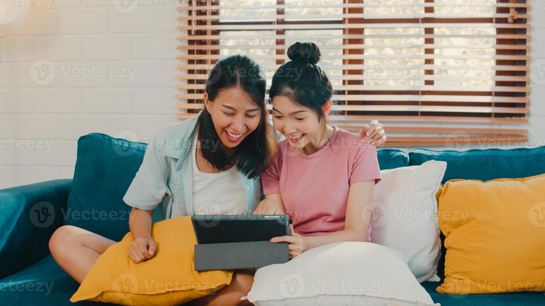 Young Lesbian lgbtq women couple using tablet at home. Asian lover female happy relax fun using technology watching movie in internet together while lying sofa in living room concept. photo