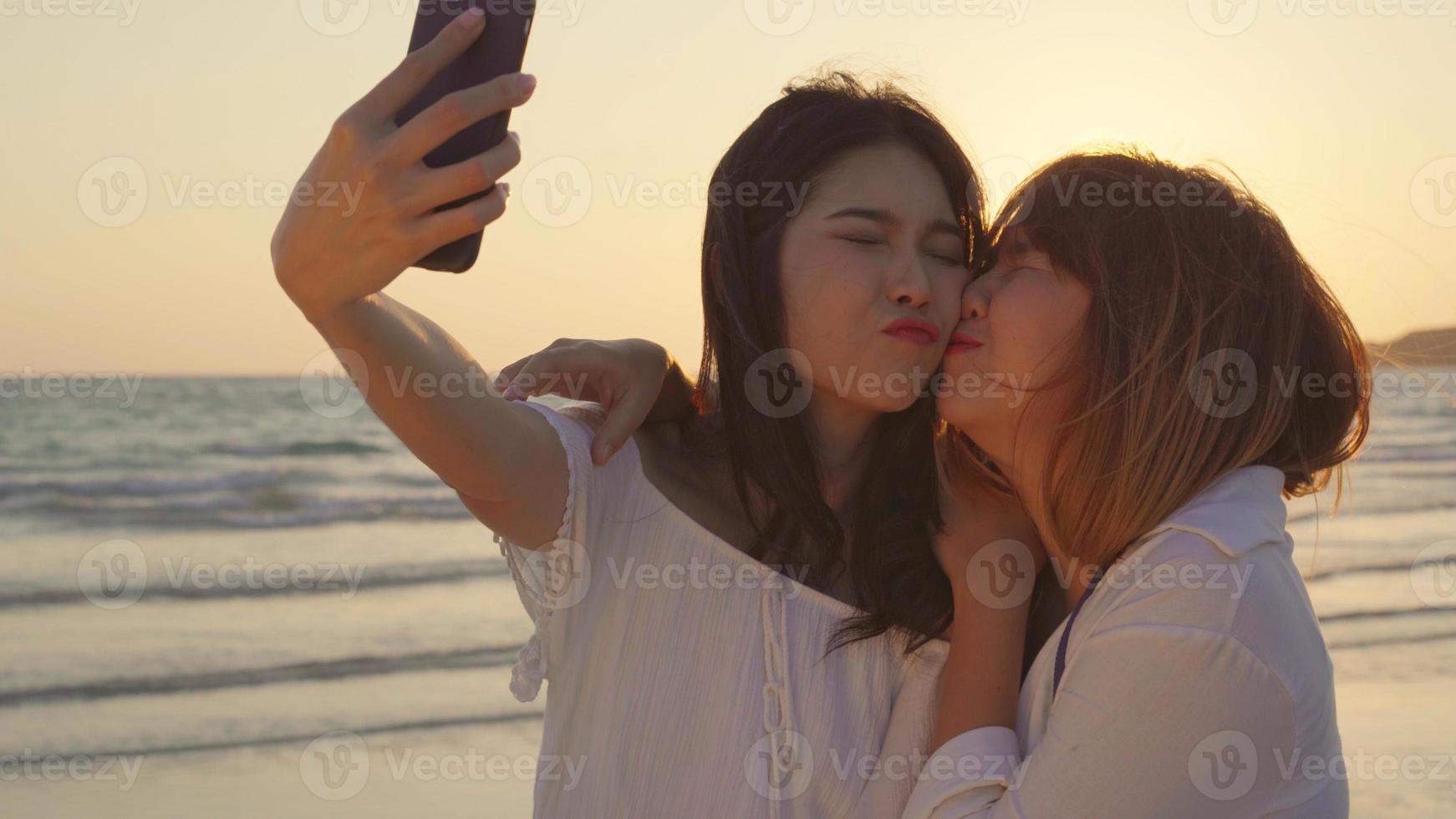 Young Asian lesbian couple using smartphone taking selfie near beach. Beautiful women lgbt couple happy relax enjoy love moment when sunset in evening. Lifestyle lesbian couple travel on beach concept photo