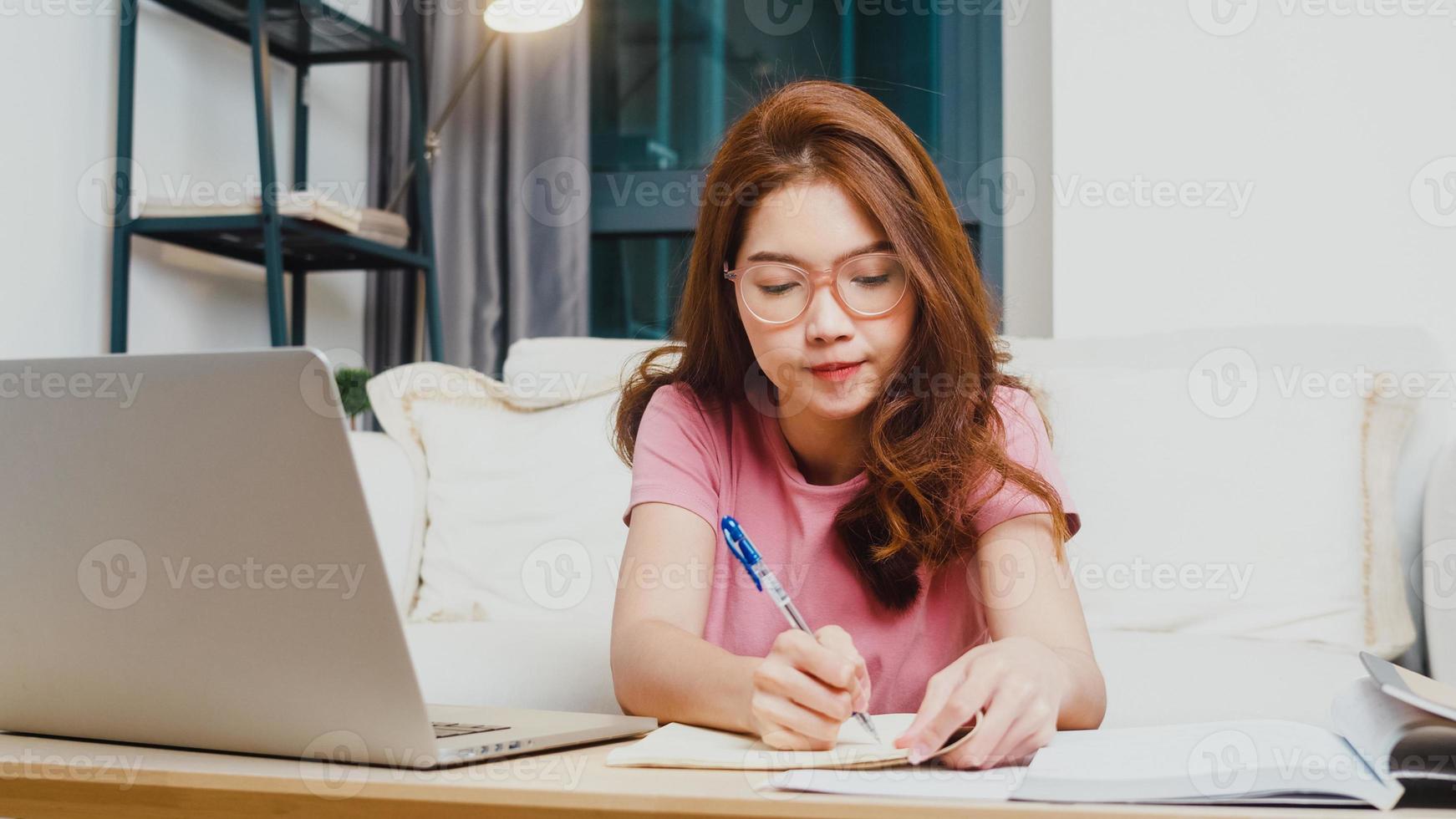 Young Asia teen girl student distance learning lesson with online teacher and study on computer laptop in living room from home at night. Social distancing, quarantine for corona virus prevention. photo