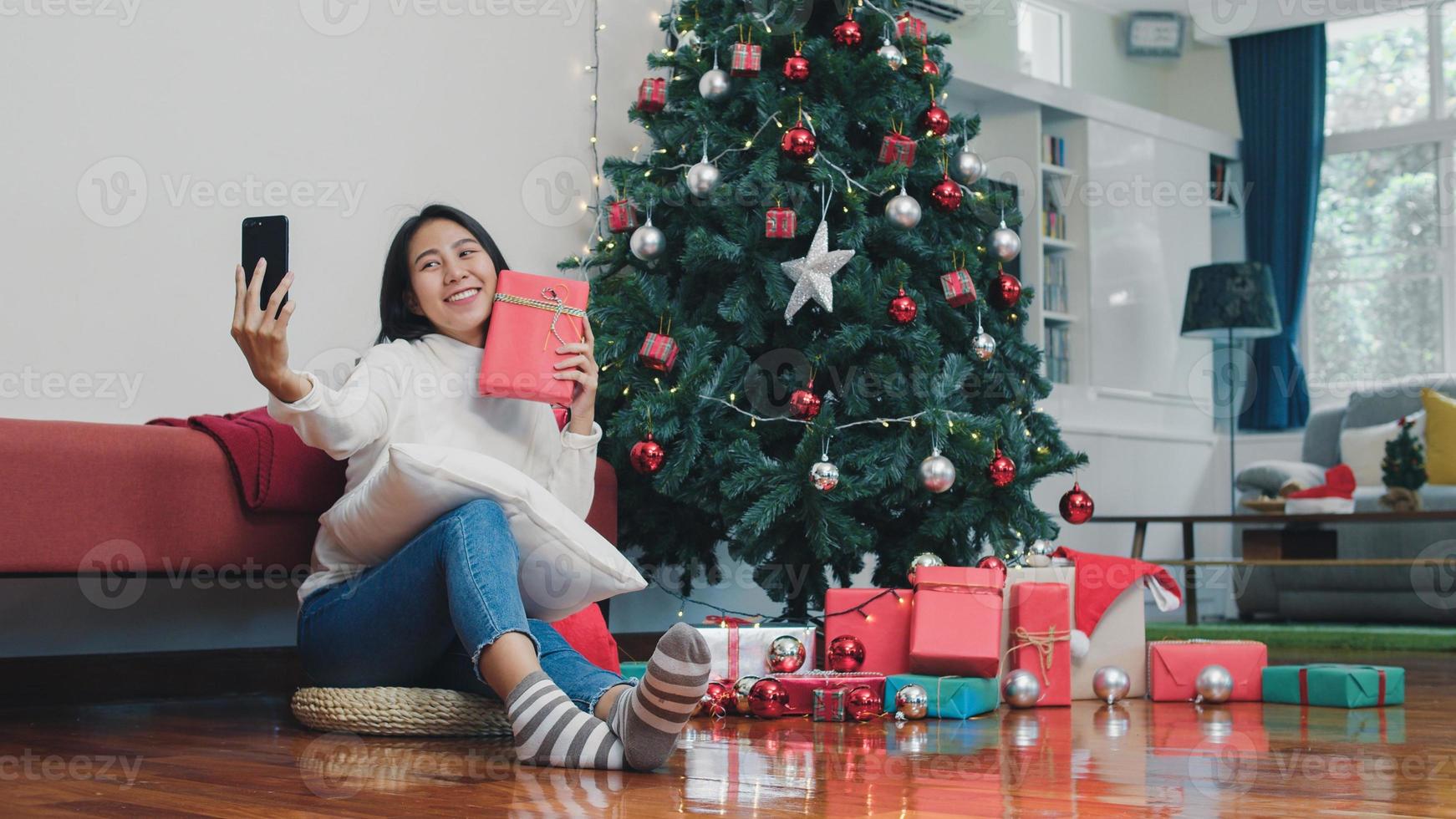 las mujeres asiáticas celebran el festival de navidad. la adolescente se relaja feliz sosteniendo un regalo y usando un smartphone selfie con un árbol de navidad disfruta de las vacaciones de invierno de navidad en la sala de estar en casa. foto