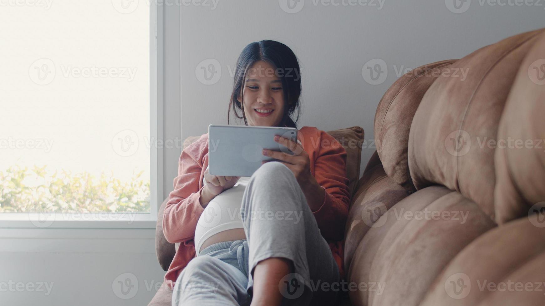 mujer embarazada asiática joven que usa la información del embarazo de la búsqueda de la tableta. mamá se siente feliz sonriendo positiva y pacífica mientras cuida a su hijo acostado en el sofá en la sala de estar en el concepto de hogar. foto