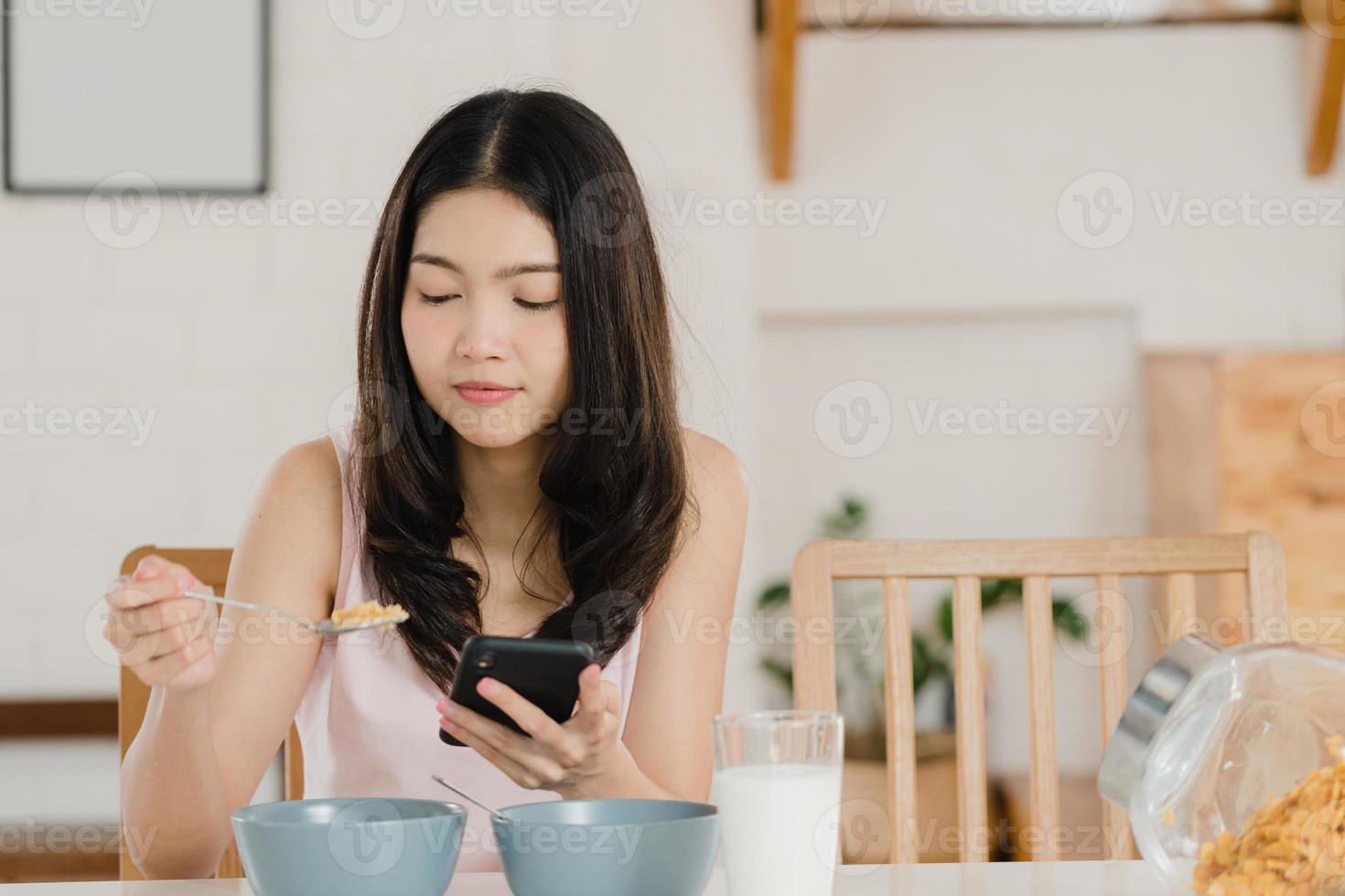 mujer japonesa asiática desayuna en casa. jóvenes asiáticas que se sienten felices usando el teléfono móvil mientras beben jugo, cereal de hojuelas de maíz y leche en un tazón en la mesa en la cocina en el concepto de la mañana. foto