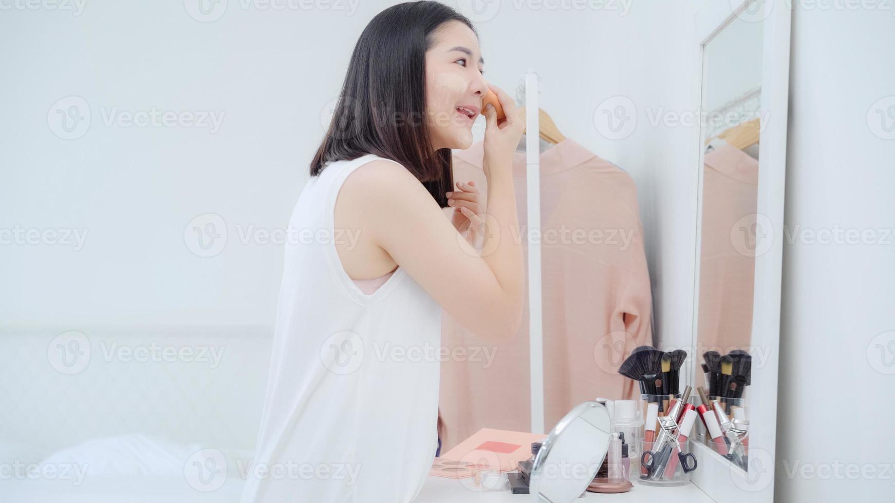 hermosa mujer asiática usando maquillaje en polvo en el espejo frontal, mujer feliz usando cosméticos de belleza para mejorar lista para trabajar en el dormitorio en casa. las mujeres de estilo de vida se relajan en el concepto de hogar. foto