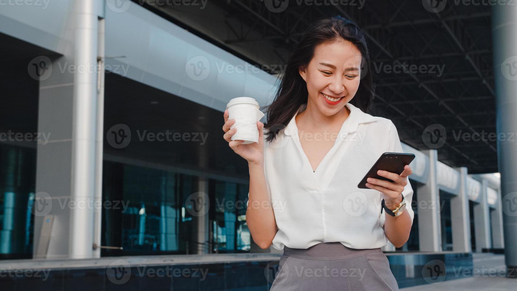 exitosa joven mujer de negocios de asia en ropa de oficina de moda sosteniendo una taza de papel desechable de bebida caliente y usando un teléfono inteligente mientras camina al aire libre en una ciudad urbana moderna. concepto de negocio sobre la marcha. foto