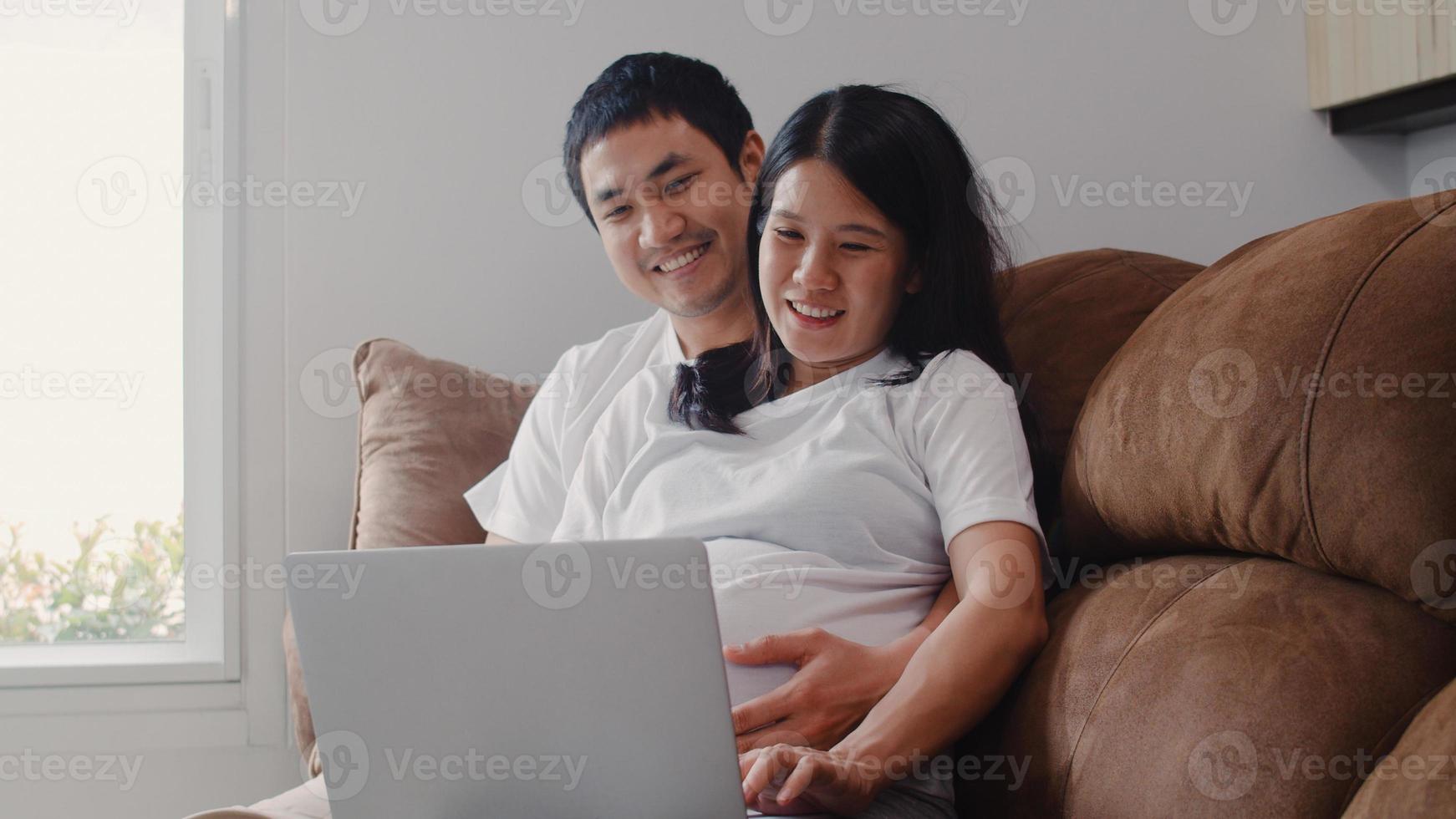 Young Asian Pregnant couple using laptop search pregnancy information. Mom and Dad feeling happy smiling positive and peaceful while take care their child lying on sofa in living room at home concept. photo