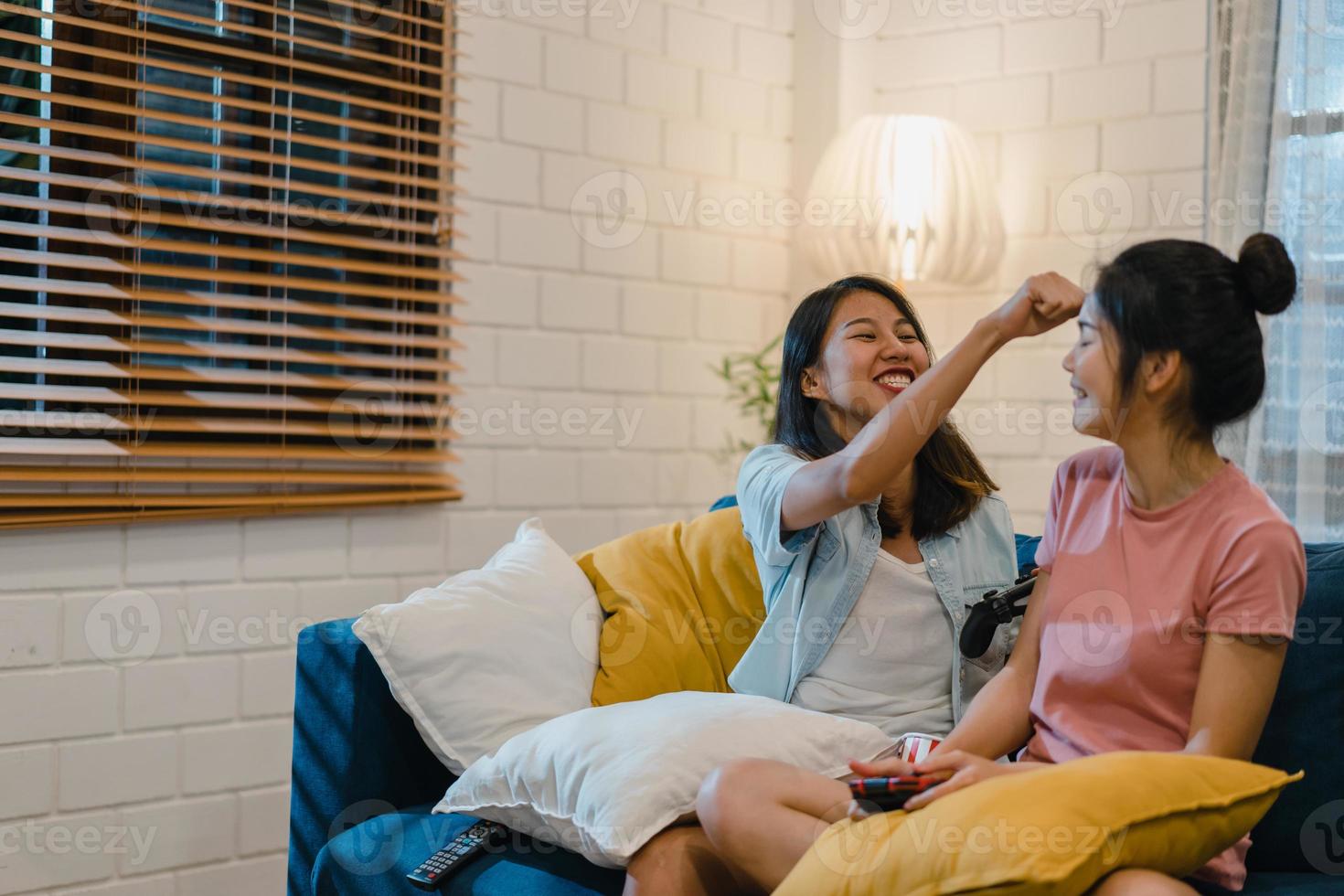 Lesbian lgbt women couple play games at home, Asian female using joystick having funny happy moment together on sofa in living room in night. Young lover football fan, celebrate holiday concept. photo