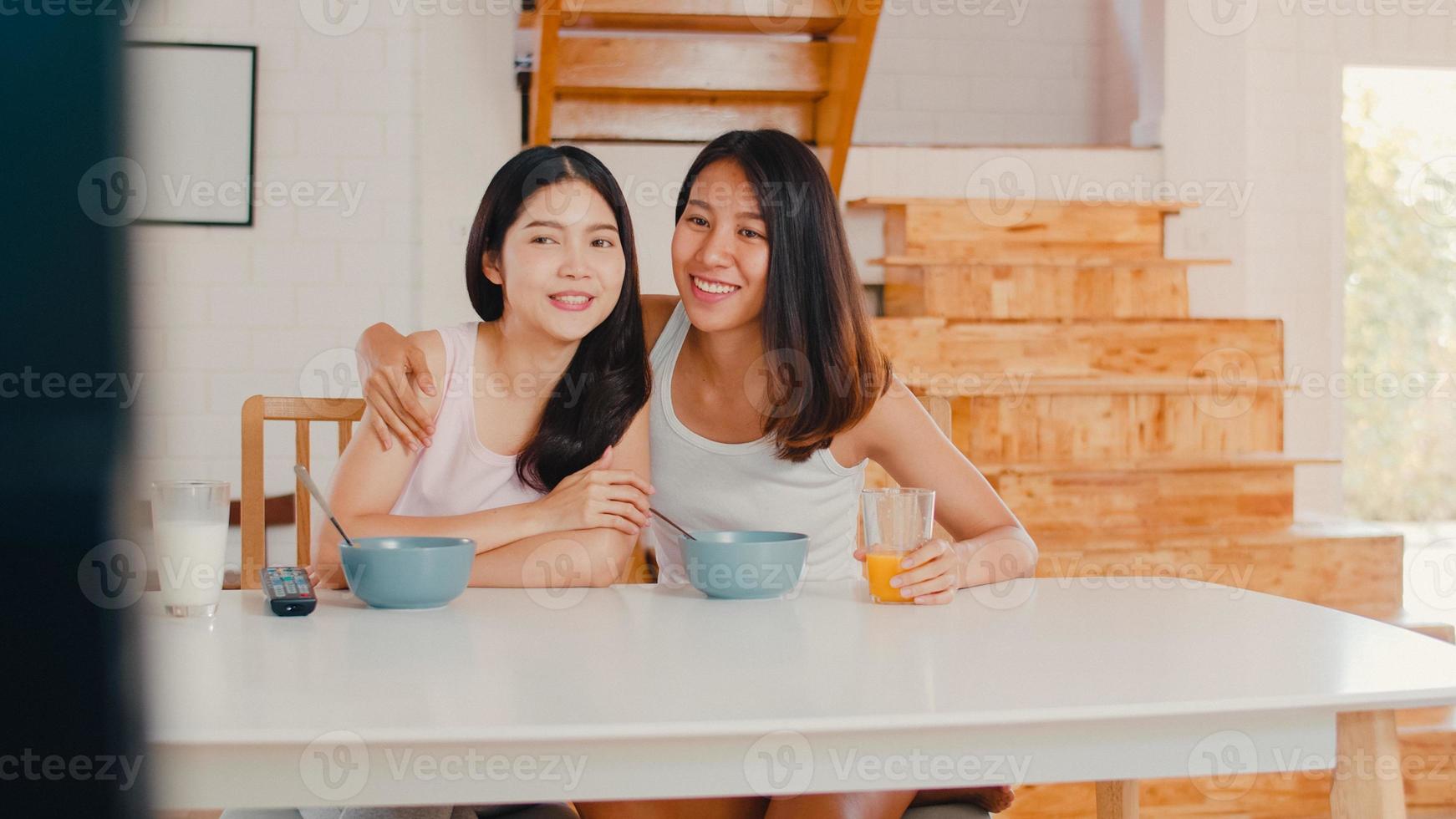 la pareja de mujeres lesbianas lgbtq asiáticas desayuna en casa, las jóvenes amantes de asia felices viendo la televisión mientras beben jugo, cereal de hojuelas de maíz y leche en un tazón en la mesa de la cocina en el concepto de la mañana. foto