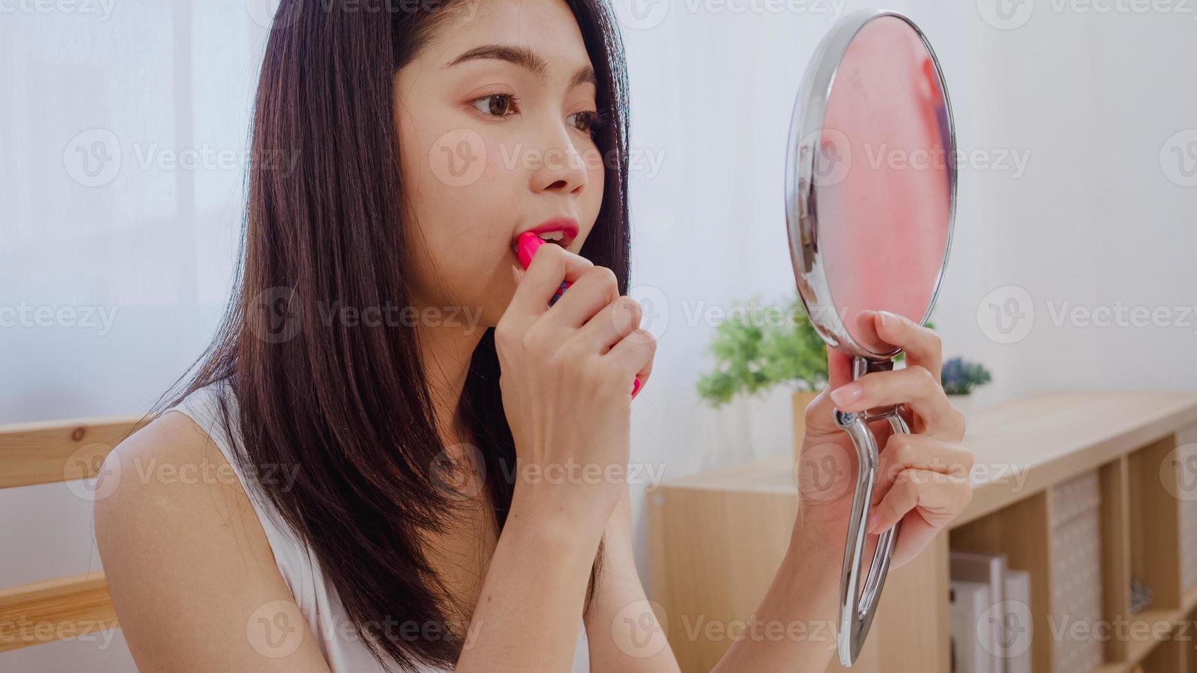 Joven asiática con maquillaje de lápiz labial frente al espejo, mujer feliz con cosméticos de belleza para mejorar, lista para trabajar en el dormitorio en casa. mujeres de estilo de vida en el concepto de hogar. foto