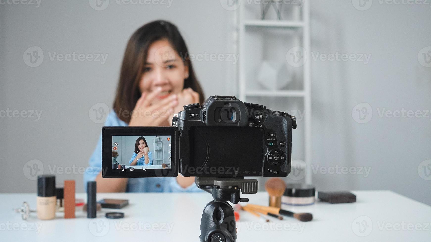 Feliz sonriente mujer asiática o blogger de belleza con pincel y cámara grabando video y agitando la mano en casa. concepto de gente de blogs de videoblog de belleza. foto