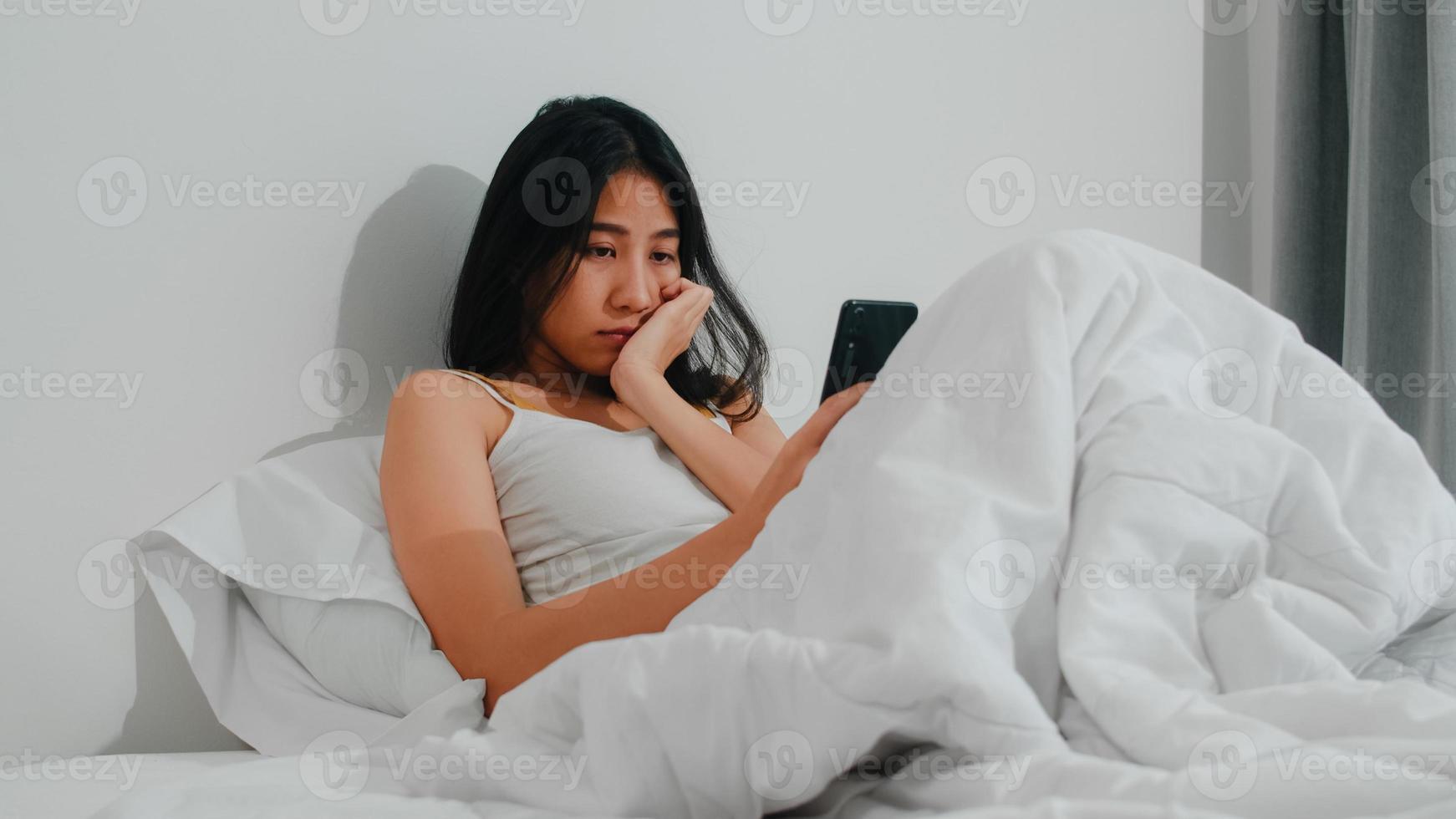 joven asiática que usa un teléfono inteligente revisando las redes sociales sintiéndose feliz sonriendo mientras está acostada en la cama después de despertarse por la mañana, hermosa y atractiva dama hispana sonriendo y relajándose en el dormitorio en casa. foto