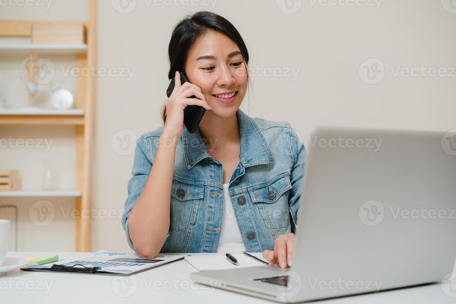 hermosa mujer asiática de negocios inteligente en ropa casual elegante trabajando en la computadora portátil y hablando por teléfono mientras está sentado en la mesa en la oficina creativa. mujeres de estilo de vida que trabajan en el concepto de hogar. foto