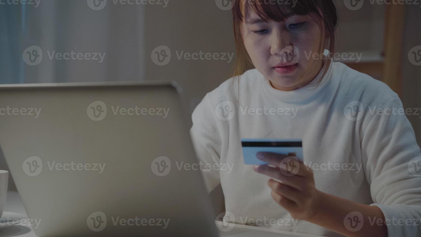 hermosa mujer asiática que usa una computadora portátil comprando compras en línea con tarjeta de crédito mientras usa ropa informal sentada en el escritorio en la sala de estar en la noche en casa. mujeres de estilo de vida que trabajan en el concepto de oficina en casa. foto