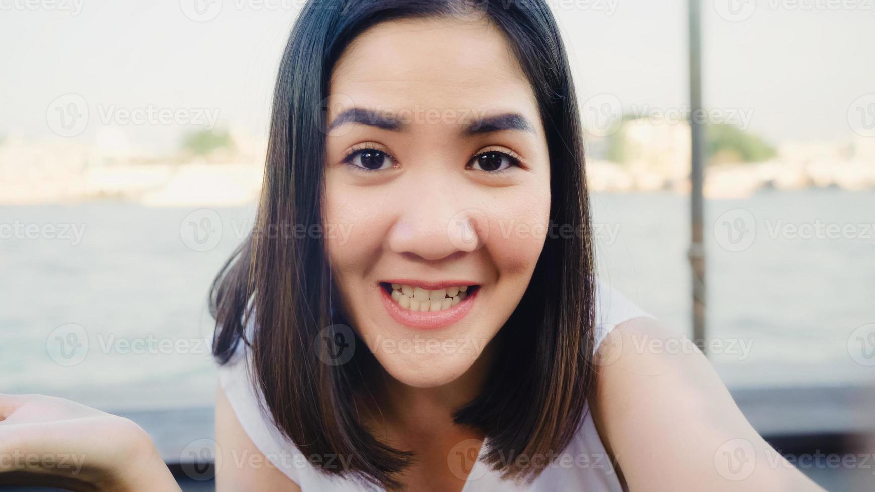 Blogger Asian friendly woman influencer looking at camera at cafe. Young lady happy relax fun using technology mobile phone record vlog video upload in social media holiday travel near river in city. photo