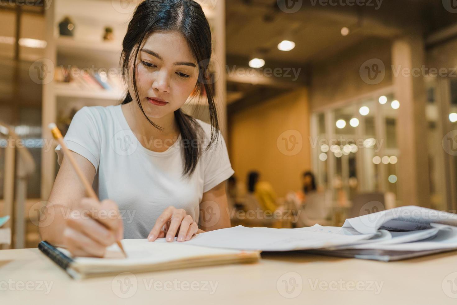 mujeres estudiantes asiáticas leyendo libros en la biblioteca de la universidad. la joven estudiante hace la tarea, lee libros de texto, estudia mucho para obtener conocimientos en el escritorio de conferencias en la noche extra del campus universitario. foto