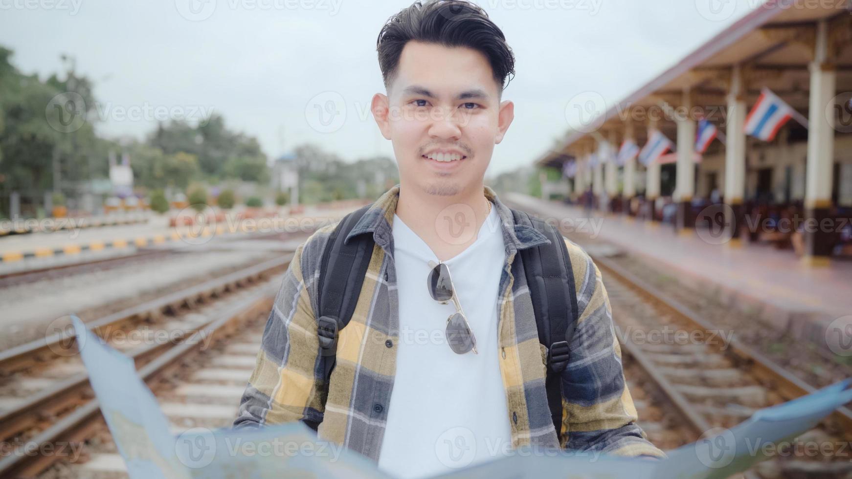 Traveler Asian man direction and looking on location map while spending holiday trip and waiting train at train station, Young male tourist backpacker enjoy journey. Lifestyle men travel concept. photo