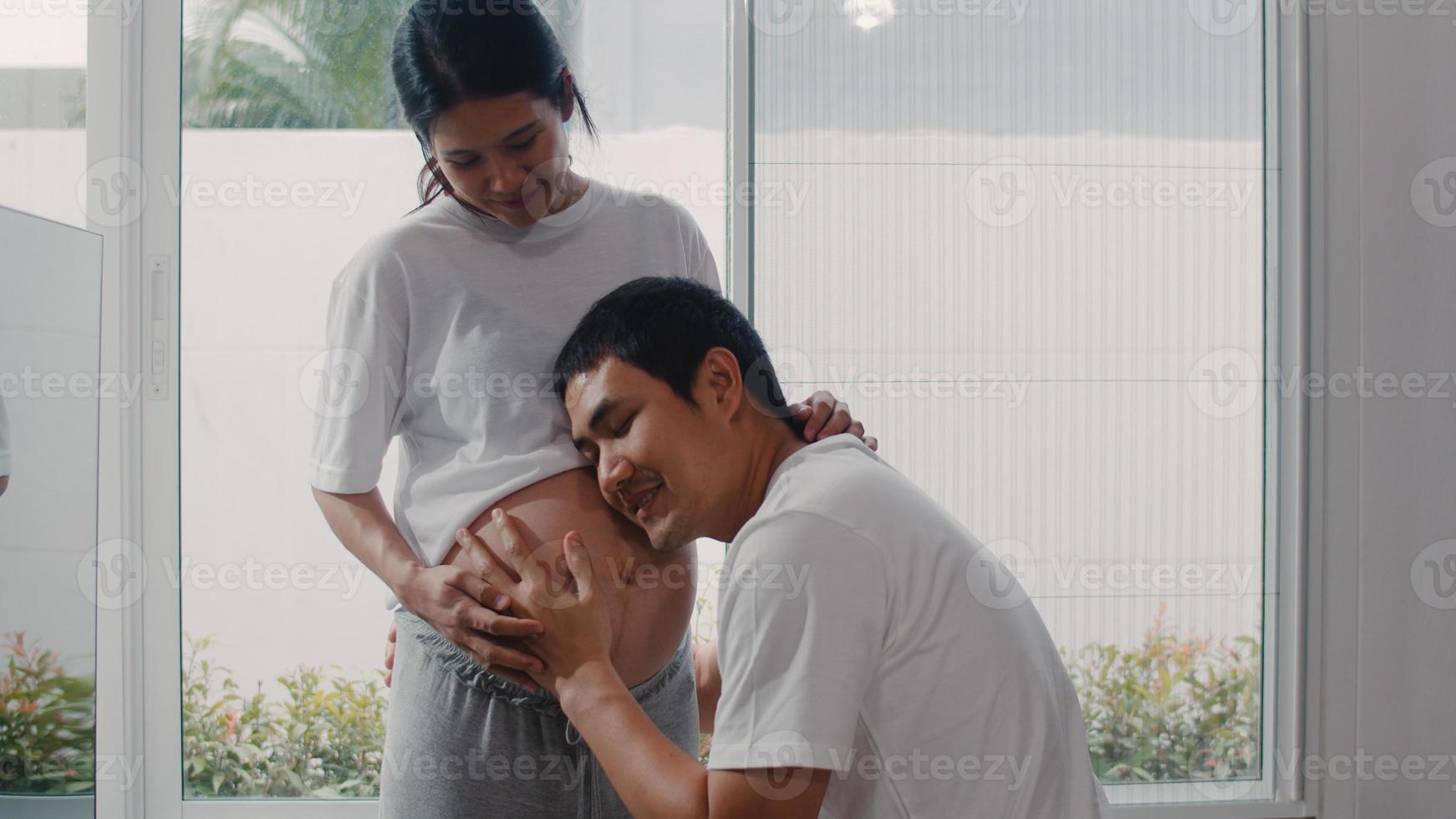 joven pareja asiática embarazada besando a su esposa en el vientre hablando con su hijo. mamá y papá se sienten felices sonriendo en paz mientras cuidan al bebé, el embarazo cerca de la ventana en la sala de estar en el concepto de hogar. foto