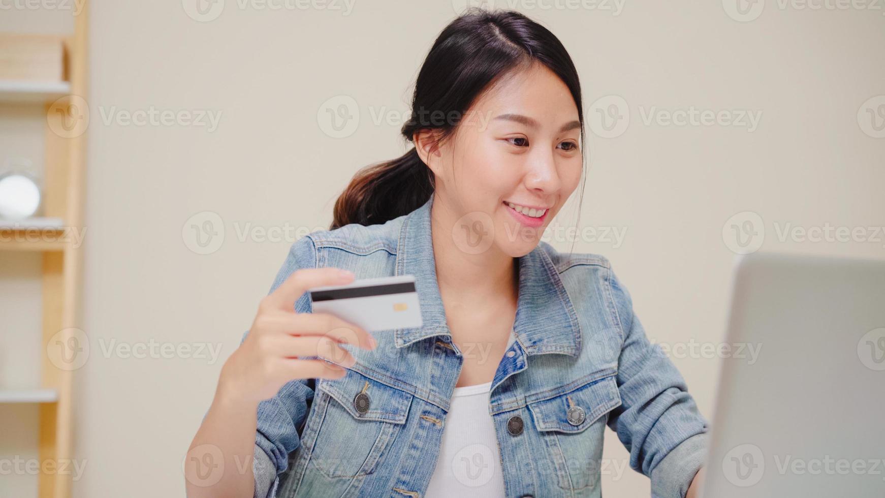 Beautiful Asian woman using computer or laptop buying online shopping by credit card while wear sweater sitting on desk in living room at home. Lifestyle woman at home concept. photo