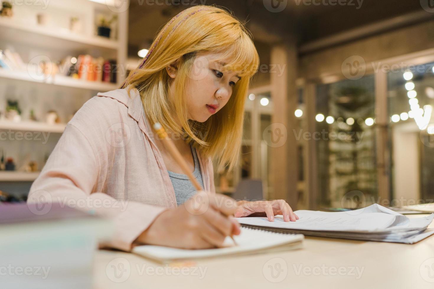 mujeres estudiantes asiáticas leyendo libros en la biblioteca de la universidad. la joven estudiante hace la tarea, lee libros de texto, estudia mucho para obtener conocimientos en el escritorio de conferencias en la noche extra del campus universitario. foto