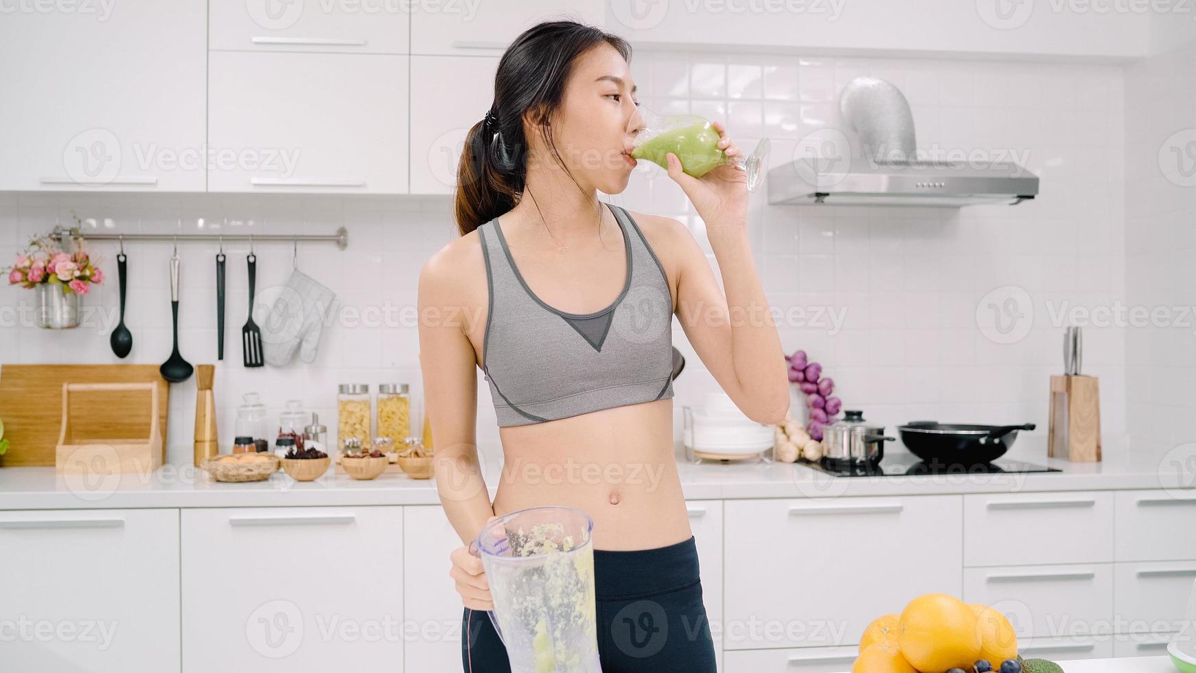 Sporty Asian woman drinking apple juice in the kitchen, beautiful female in sport clothing use organic fruits lots of nutrition making apple juice by herself at home. Healthy food concept. photo