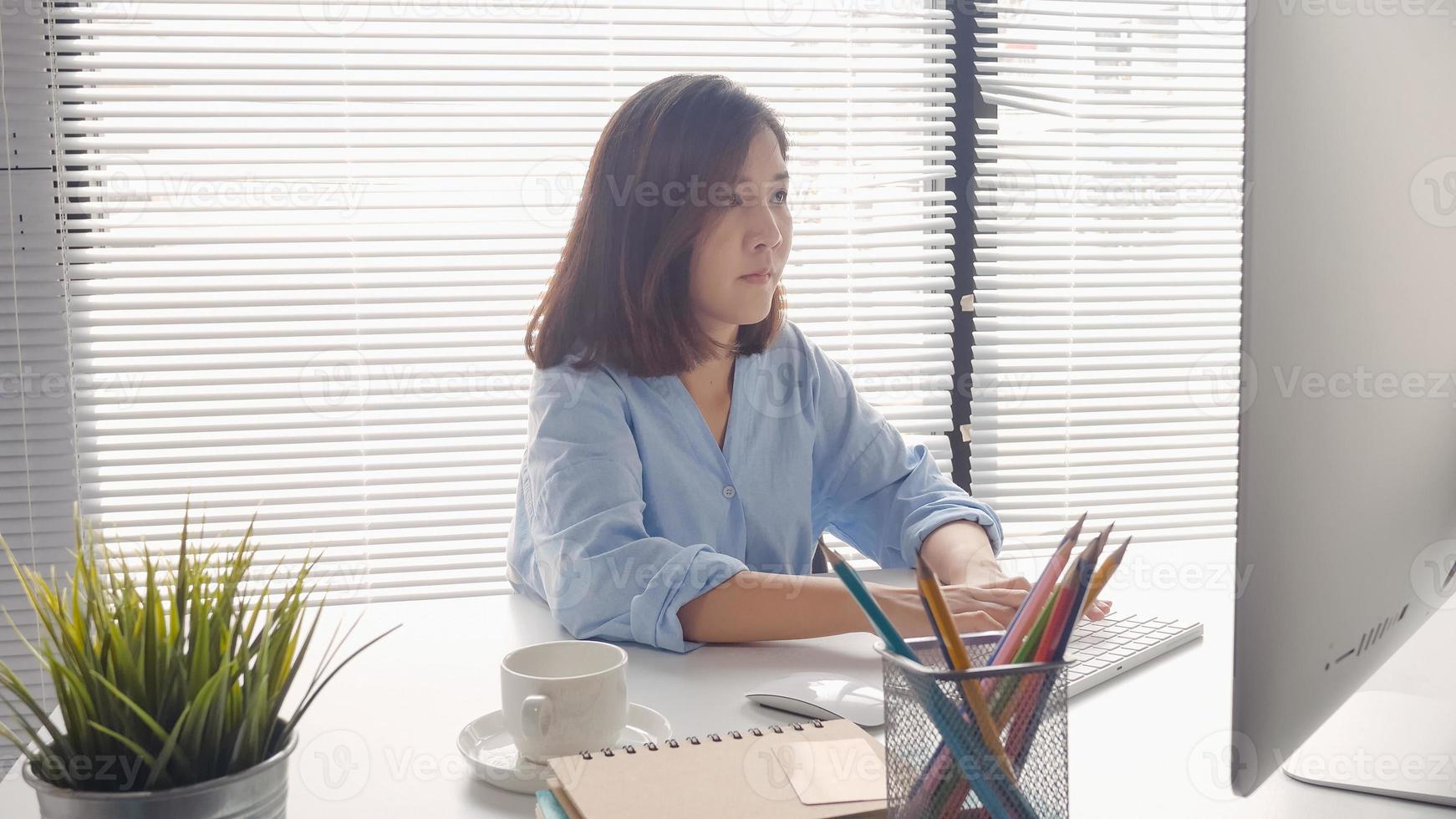 Beautiful young smiling Asian woman working on laptop while at home in office work space. Businesswoman working from home via portable computer writing on keyboard. Enjoying time at home. photo