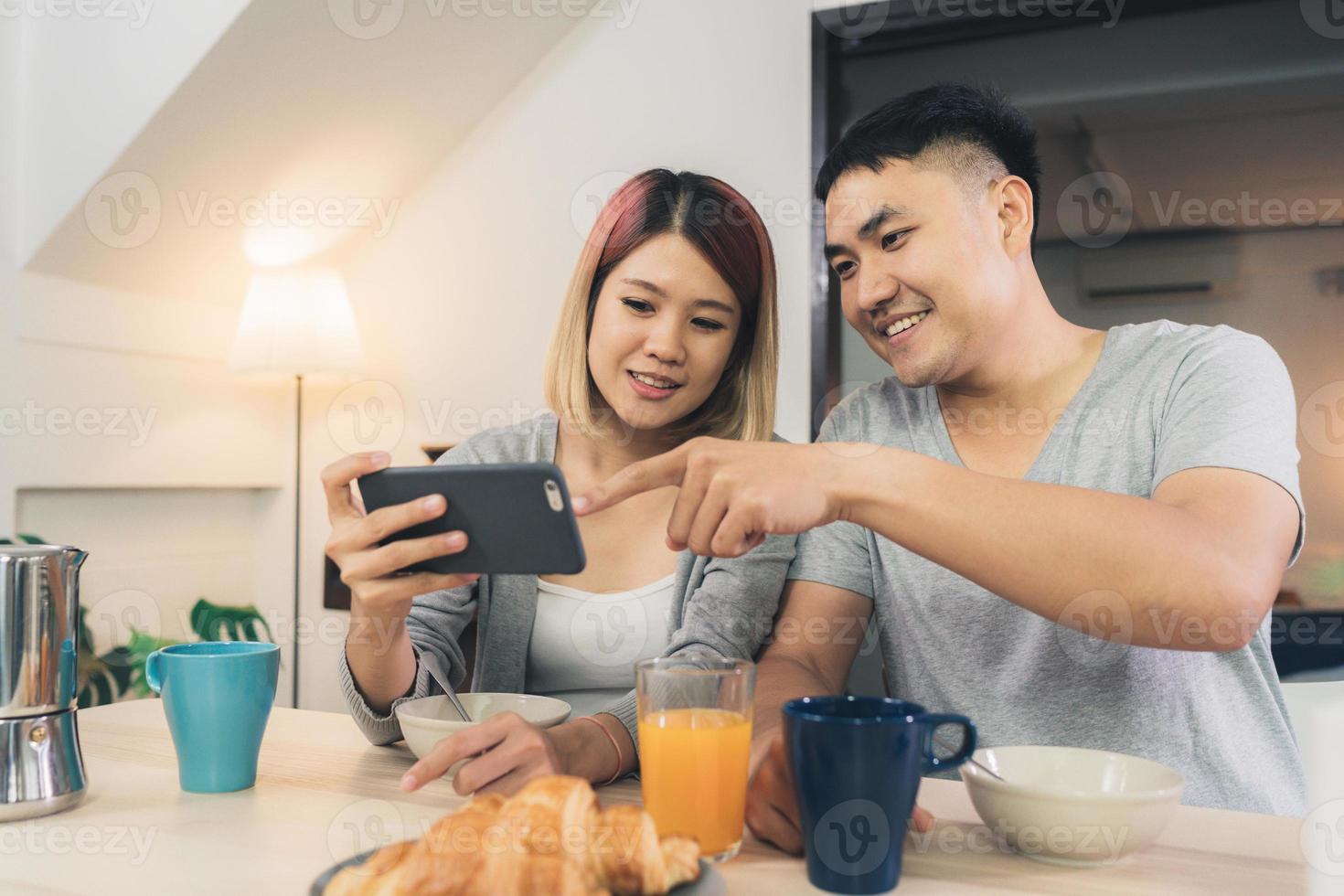 Atractiva pareja asiática joven distraída en la mesa con el periódico y el teléfono celular mientras desayuna. Emocionada joven pareja asiática sorprendida por una increíble buena noticia, familia feliz asombrada por Internet. foto