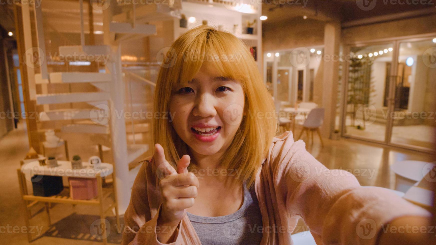 Blogger Asian friendly women influencer waving hand looking at camera at night cafe. Young lady happy relax fun using technology mobile phone record vlog video upload in social media at college campus photo