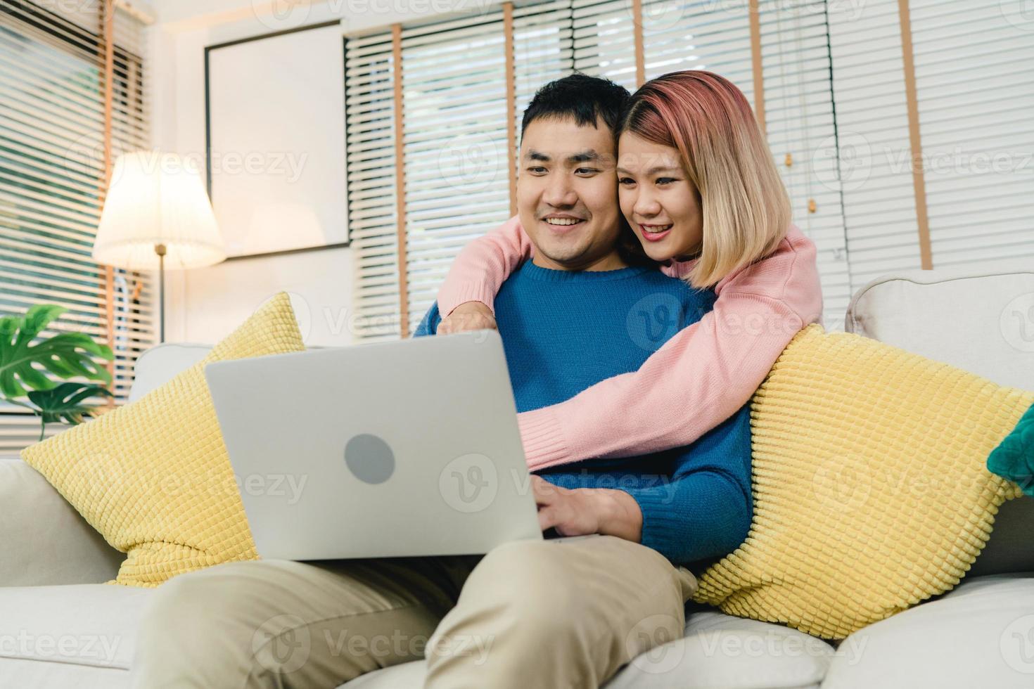 atractiva pareja dulce asiática que usa computadora o computadora portátil mientras está acostado en el sofá cuando se relaja en su sala de estar en casa. el esposo y su esposa usan el concepto de relajación y tiempo romántico en casa. foto