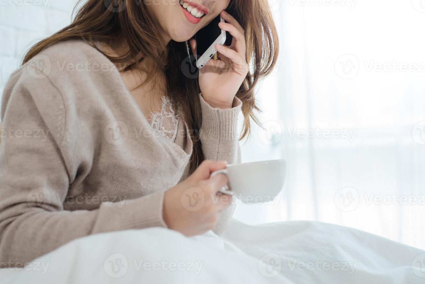 Asian woman using the smartphone on her bed while holding cup of coffee in the morning. Beautiful asian woman enjoying warm coffee and talking on telephone in her bedroom. lifestyle asia woman concept photo