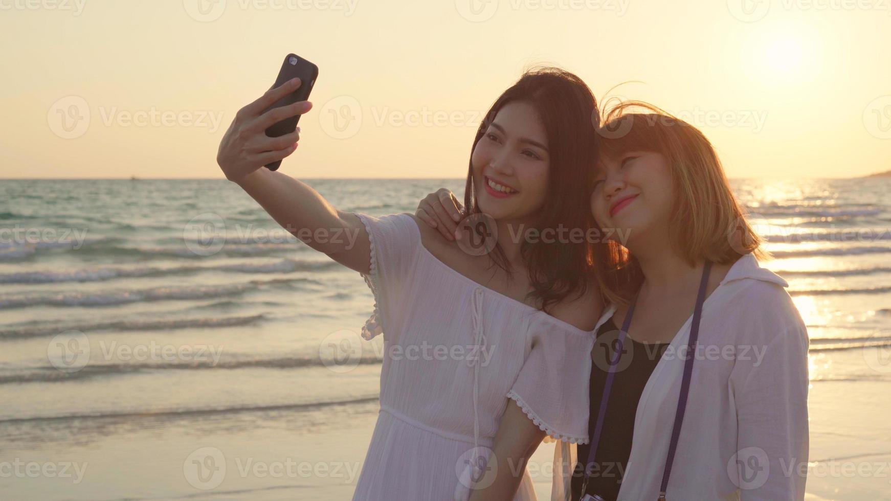 Young Asian lesbian couple using smartphone taking selfie near beach. Beautiful women lgbt couple happy relax enjoy love moment when sunset in evening. Lifestyle lesbian couple travel on beach concept photo