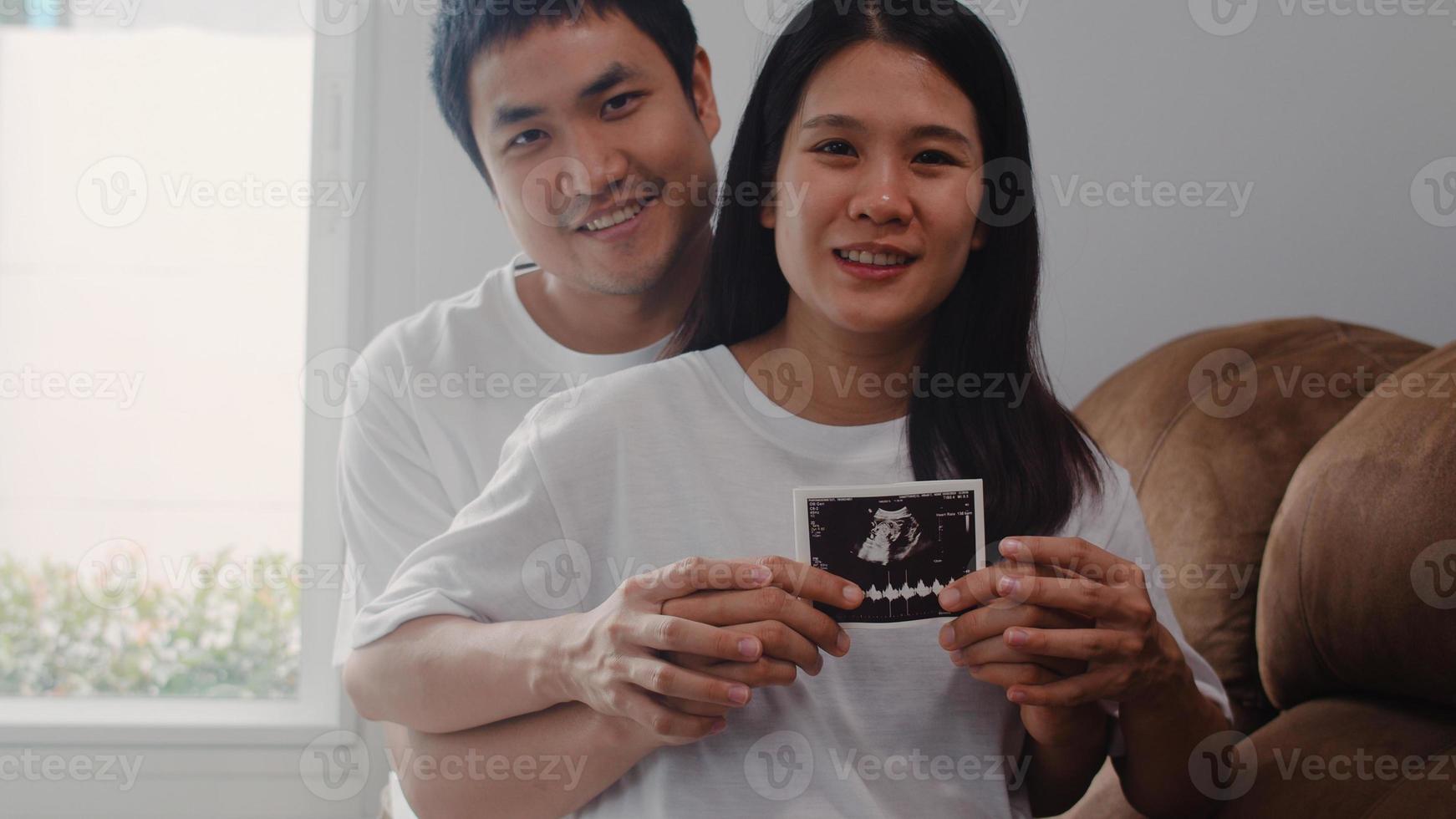 Young Asian Pregnant couple show and looking ultrasound photo baby in belly. Mom and Dad feeling happy smiling peaceful while take care child lying on sofa in living room at home concept.