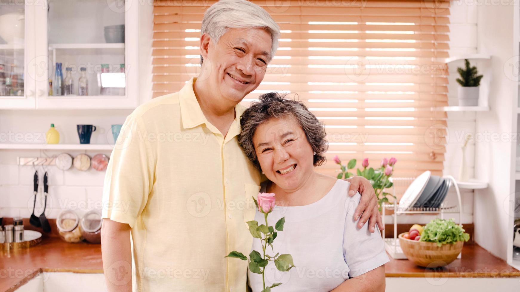 Asian elderly couple feeling happy smiling and holding flower and looking to camera while relax in kitchen at home. Lifestyle Senior family enjoy time at home concept. Portrait looking at camera. photo