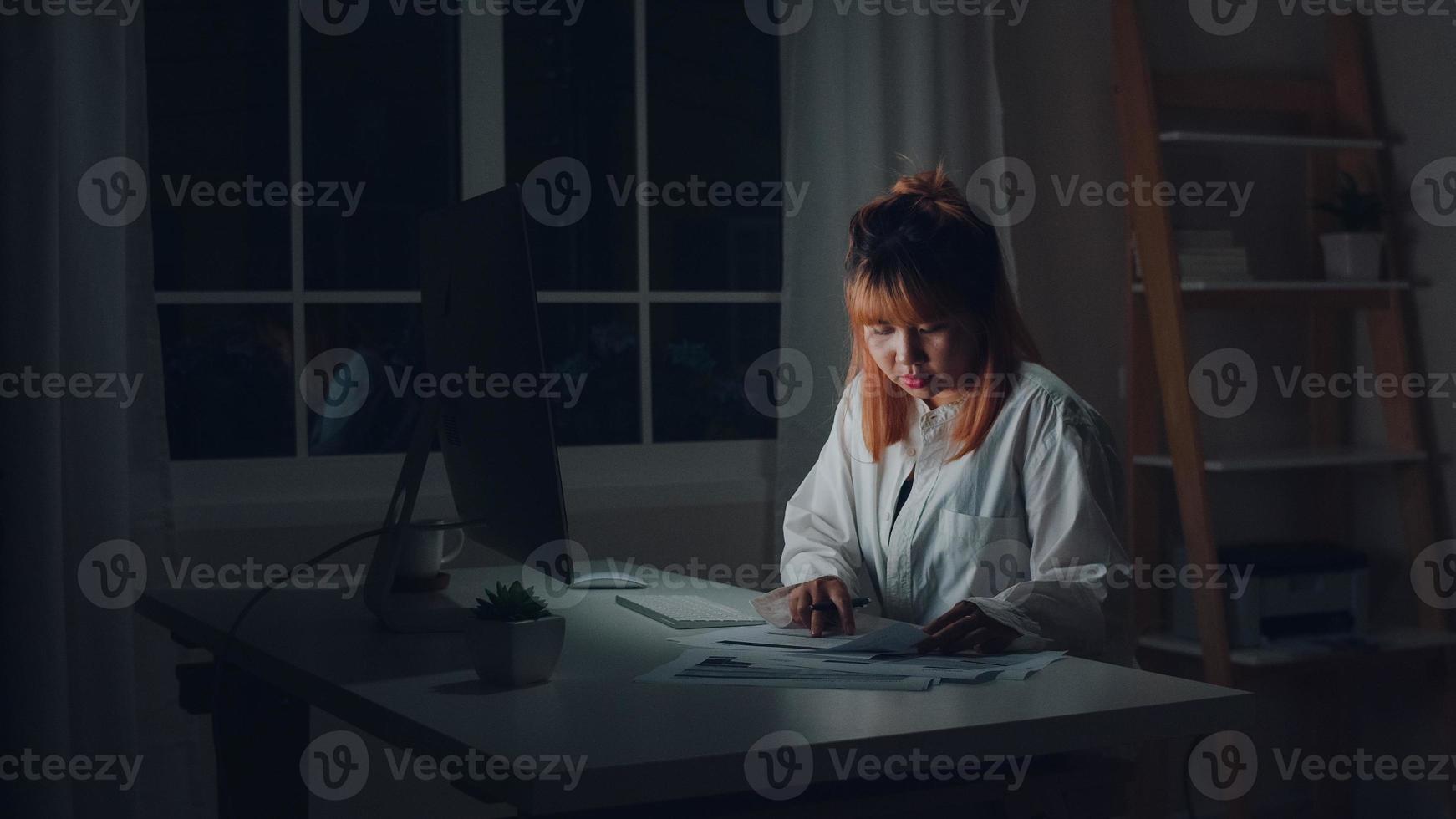 Young asian woman working late using desktop on desk in living room at home. Asia business woman writing notebook document finance and calculator in night at home office. Overworked female concept. photo