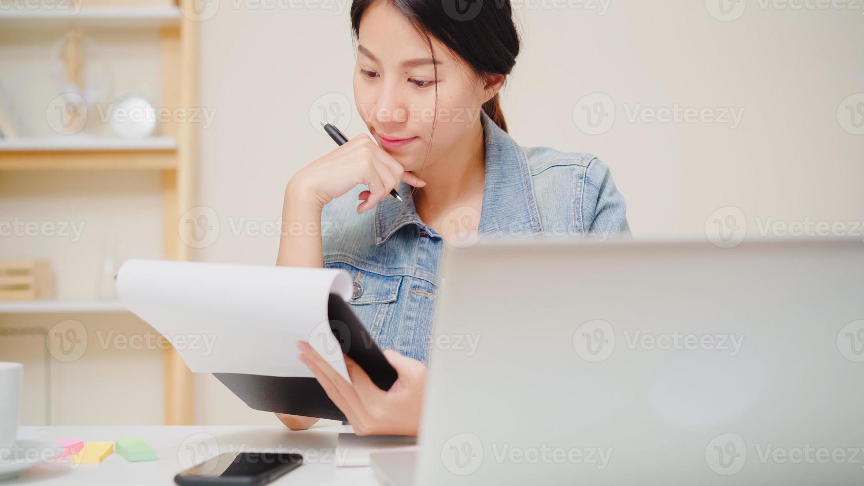 Beautiful young smiling asian woman working laptop on desk in living room at home. Asia business woman writing notebook document finance and calculator in home office. Enjoying time at home concept. photo