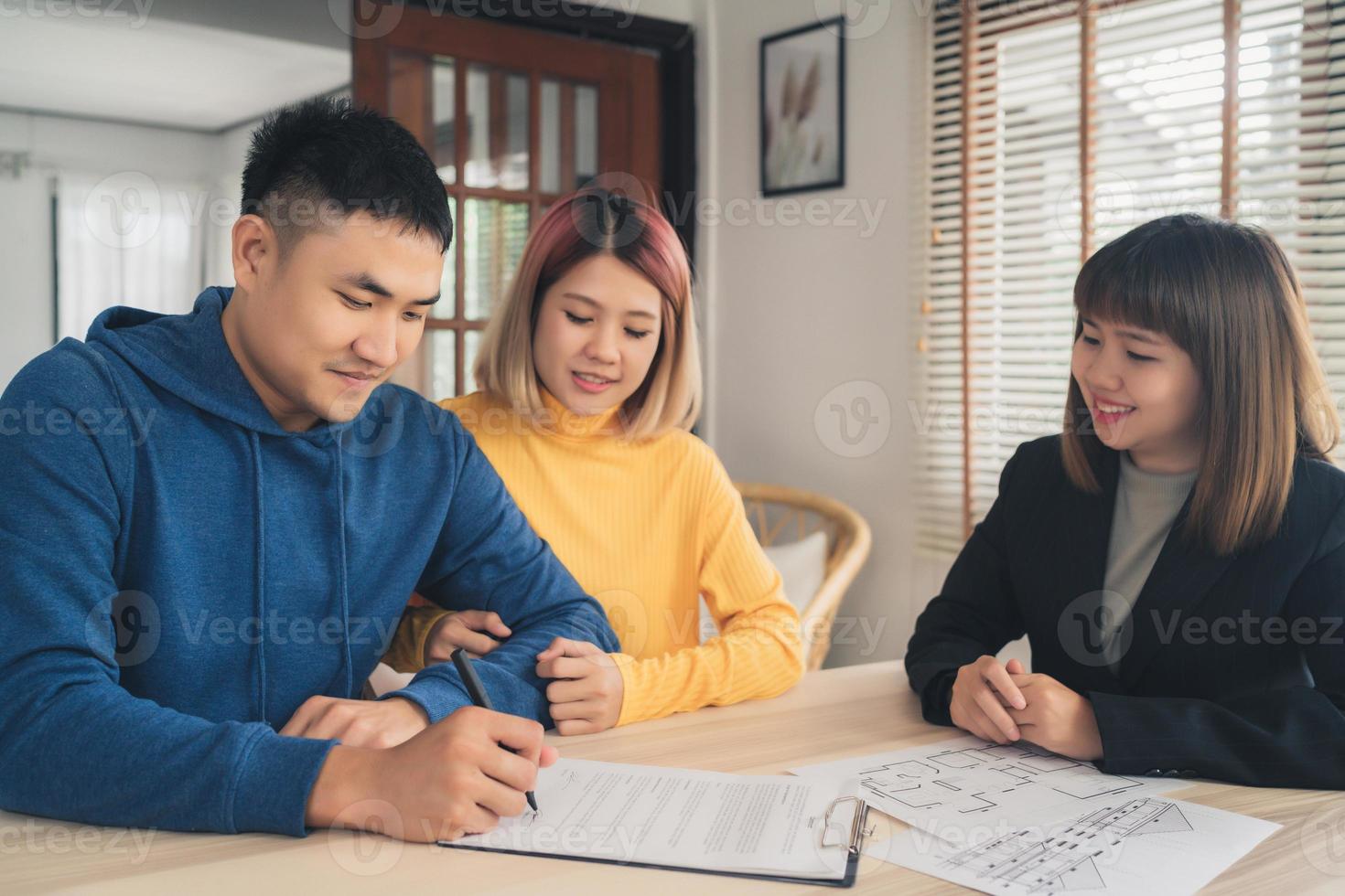 Happy young Asian couple and realtor agent. Cheerful young man signing some documents while sitting at desk together with his wife. Buying new house real estate. Signing good condition contract. photo