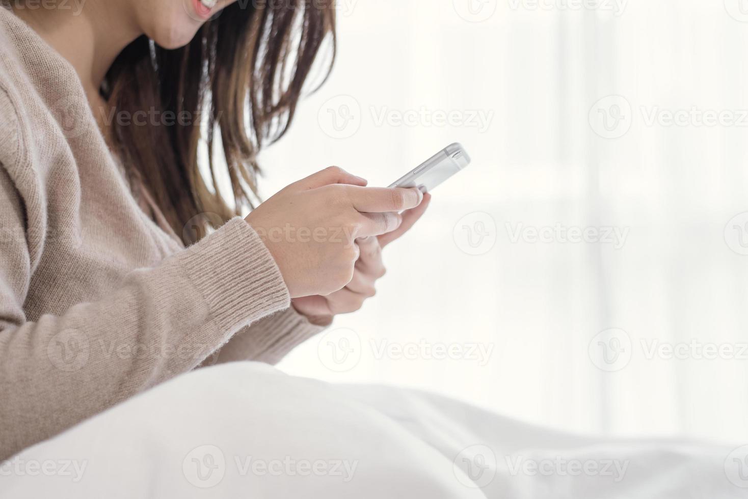 Las mujeres asiáticas felices están usando teléfonos inteligentes en la cama por la mañana. mujer asiática en la cama comprobando aplicaciones sociales con smartphone. mujer sonriente navegando por la red con el teléfono móvil en casa. concepto de adicto al móvil. foto