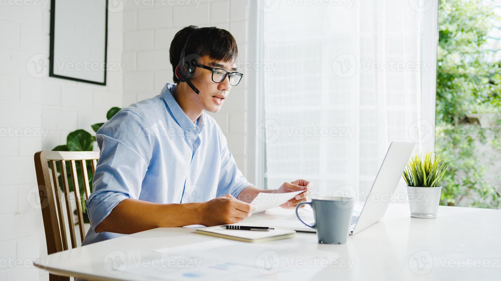 Young Asia businessman using laptop talk to colleagues about plan in video call while smart working from home at living room. Self-isolation, social distancing, quarantine for corona virus prevention. photo