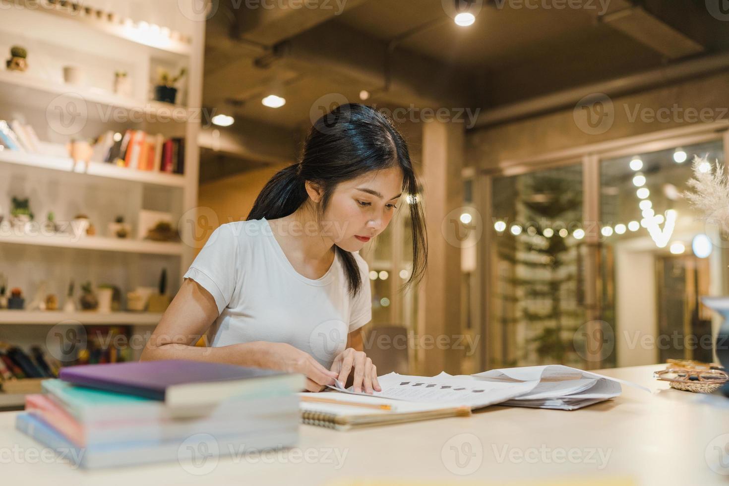 mujeres estudiantes asiáticas leyendo libros en la biblioteca de la universidad. la joven estudiante hace la tarea, lee libros de texto, estudia mucho para obtener conocimientos en el escritorio de conferencias en la noche extra del campus universitario. foto