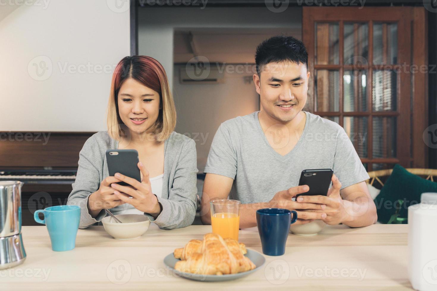 Atractiva pareja asiática joven distraída en la mesa con el periódico y el teléfono celular mientras desayuna. Emocionada joven pareja asiática sorprendida por una increíble buena noticia, familia feliz asombrada por Internet. foto