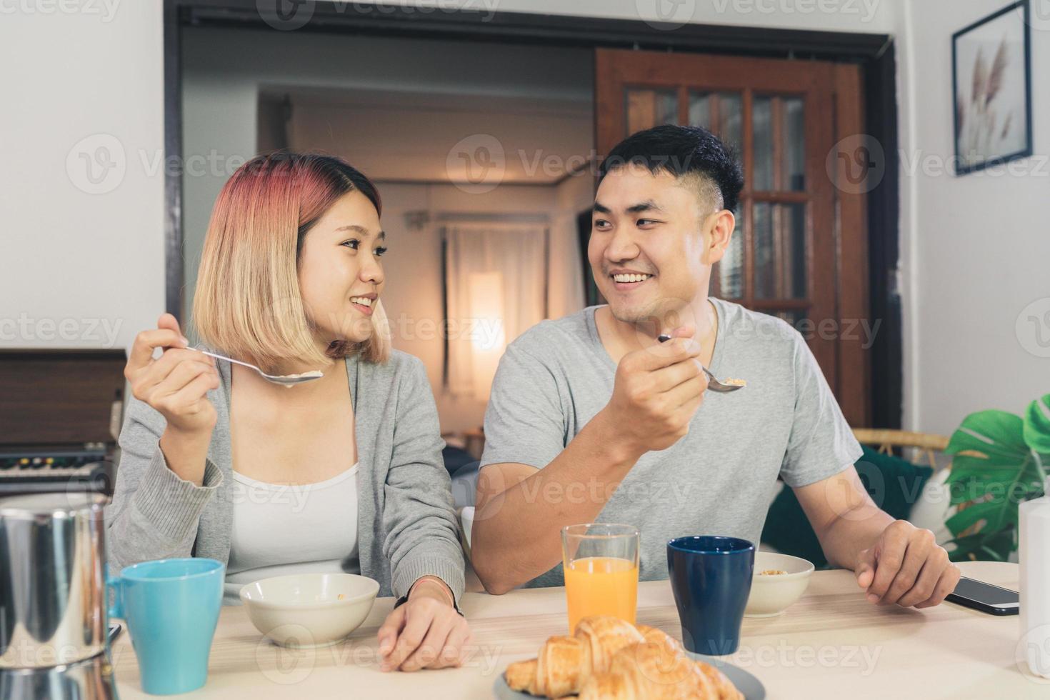 Happy sweet Asian couple having breakfast, cereal in milk, bread and drinking orange juice after wake up in the morning. Husband and his wife eating food together. photo