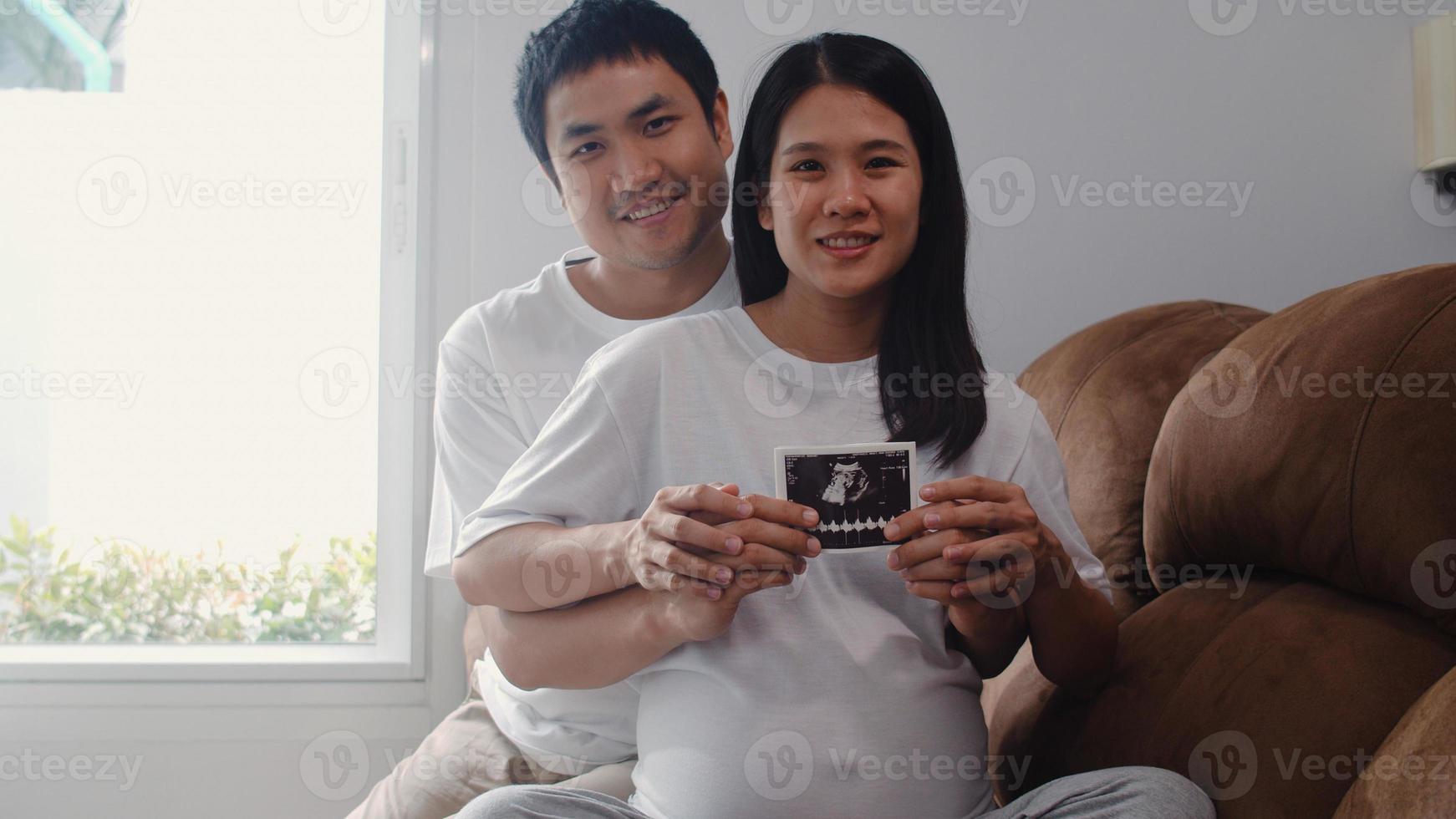 Young Asian Pregnant couple show and looking ultrasound photo baby in belly. Mom and Dad feeling happy smiling peaceful while take care child lying on sofa in living room at home concept.