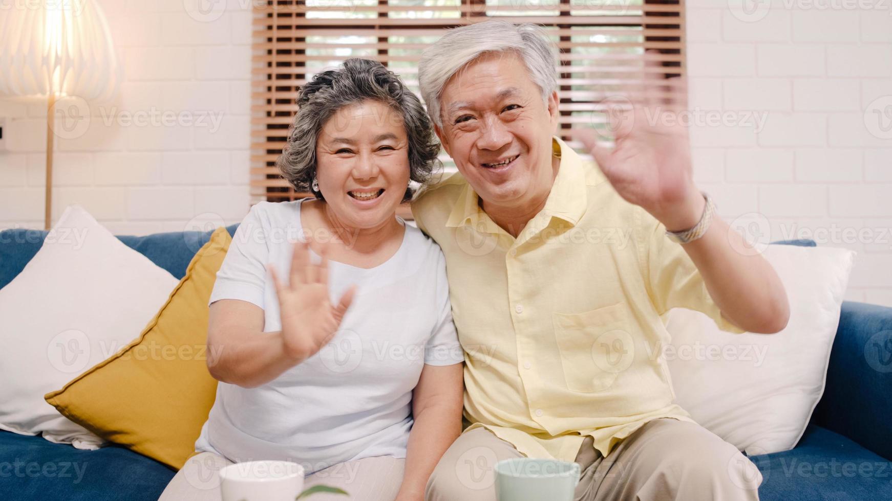 Asian elderly couple using smartphone video conference with grandchild while lying on sofa in living room at home. Enjoying time lifestyle senior family at home concept. Portrait looking at camera. photo