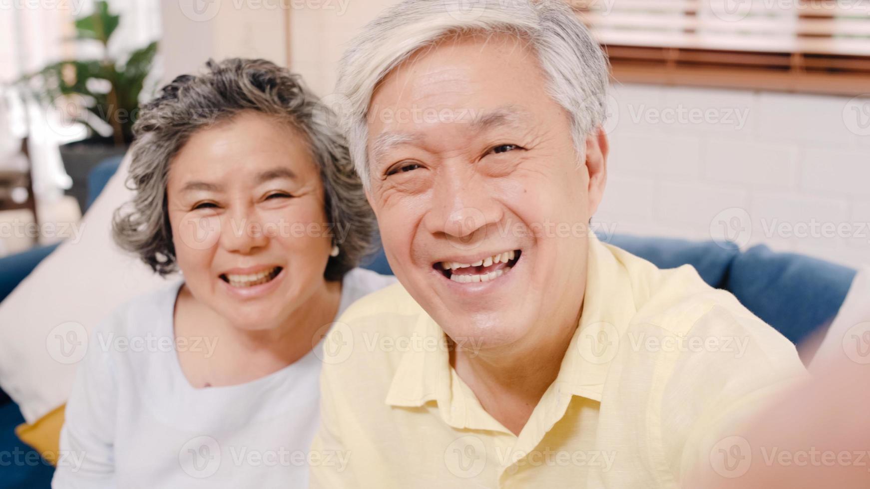 Asian elderly couple using smartphone video conference with grandchild while lying on sofa in living room at home. Enjoying time lifestyle senior family at home concept. Portrait looking at camera. photo